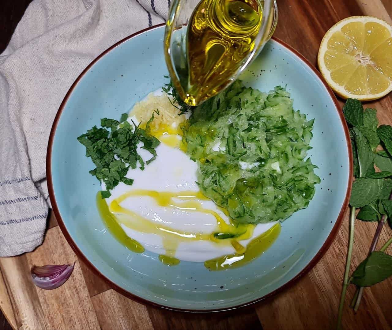 Ingredients for vegan tzatziki sauce in a bowl, including grated cucumber, chopped mint, minced garlic, and olive oil being poured.