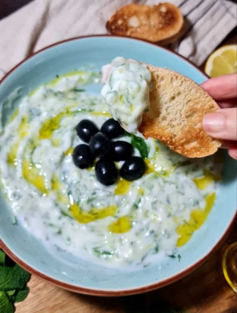 A bowl of vegan tzatziki sauce garnished with black olives and a drizzle of olive oil, with a hand holding a piece of toasted bread dipped in the sauce.