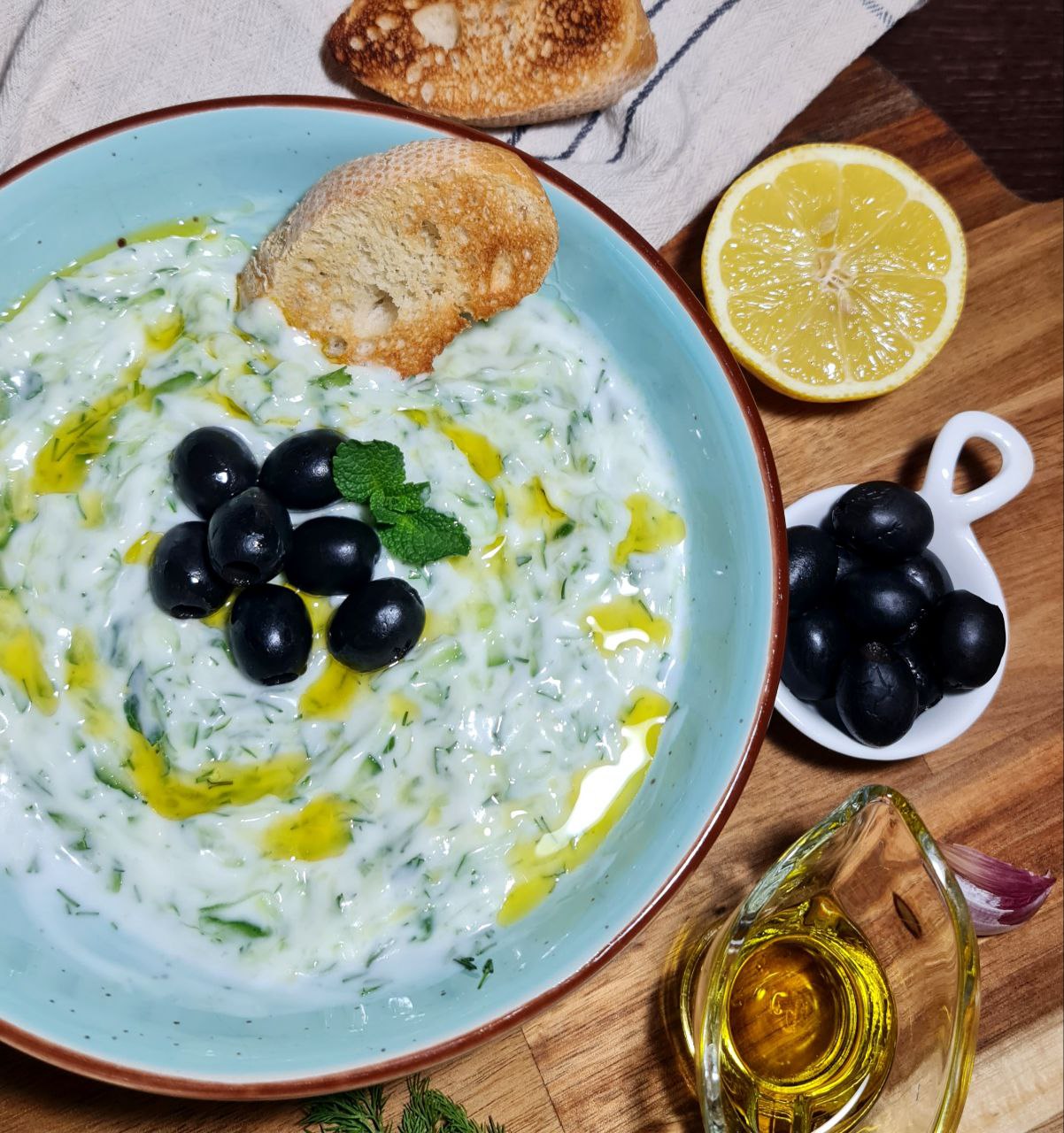 A bowl of vegan tzatziki sauce garnished with black olives, mint leaves, and a drizzle of olive oil, served with toasted bread and a lemon half on the side.