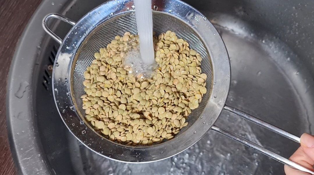 A person rinsing lentils in a metal strainer under running water in a kitchen sink.