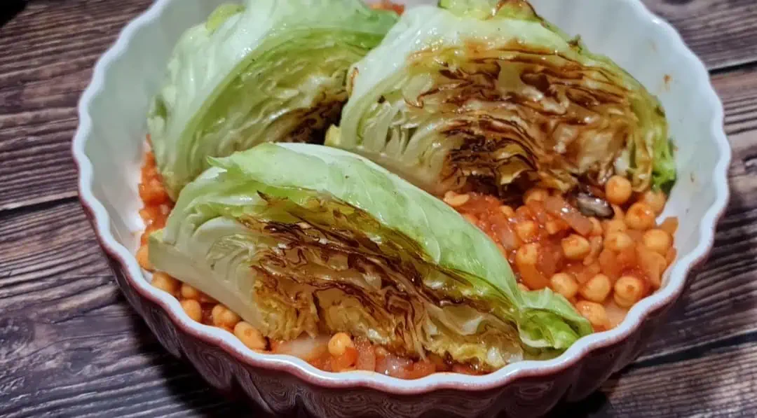 A dish consisting of three wedges of grilled or roasted cabbage placed on top of a bed of small, round pasta pieces in a tomato-based sauce, presented in a white, scalloped-edged bowl on a wooden surface.