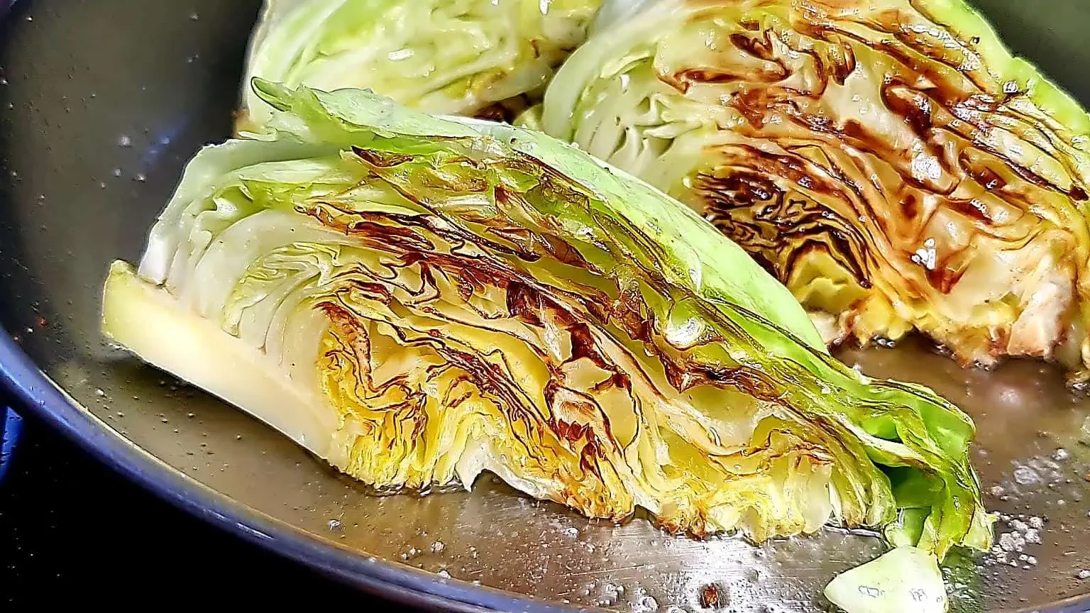 A close-up of a pan containing wedges of cabbage that have been grilled. The cabbage wedges have visible char marks and a slightly browned appearance, with outer leaves green and inner layers a mix of pale green and yellow.