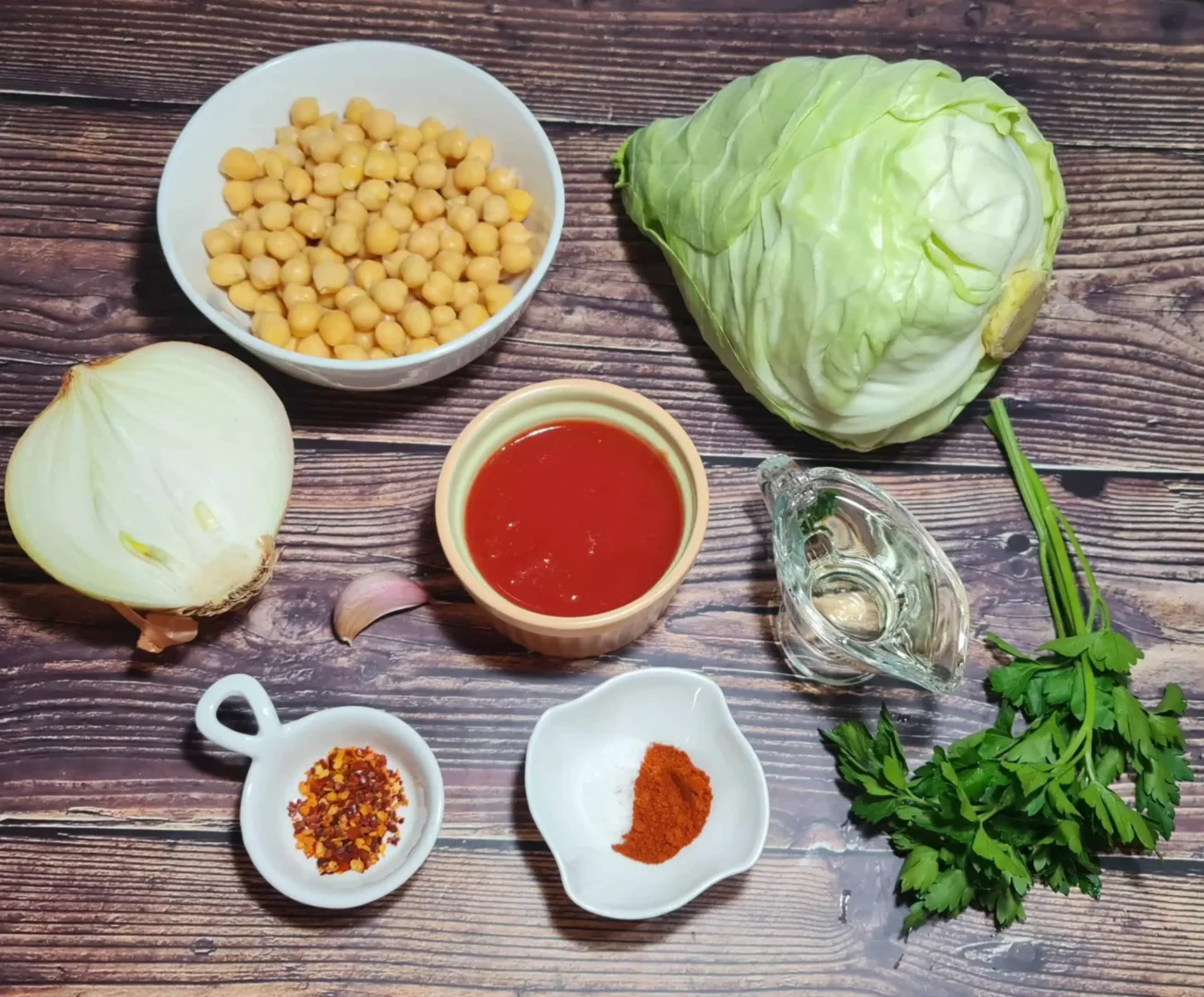 Ingredients for preparing Cabbage Wedges with Chickpeas, including a bowl of chickpeas, a whole cabbage, half a white onion, a clove of garlic, tomato sauce, red pepper flakes, paprika, oil, and fresh parsley.