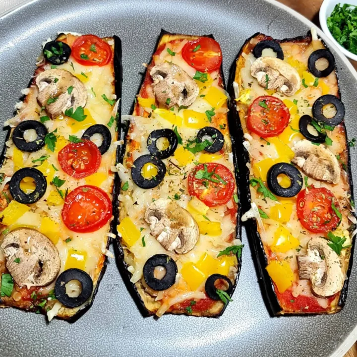 I could eat these delicious eggplants every day! Very quick and easy❗️ Three slices of eggplant prepared as mini pizzas, each topped with cherry tomato halves, black olive slices, yellow bell pepper pieces, mushroom slices, shredded cheese, and chopped parsley, arranged on a gray plate with a background of fresh cherry tomatoes on the vine, a dish with chopped herbs, and a striped cloth napkin.