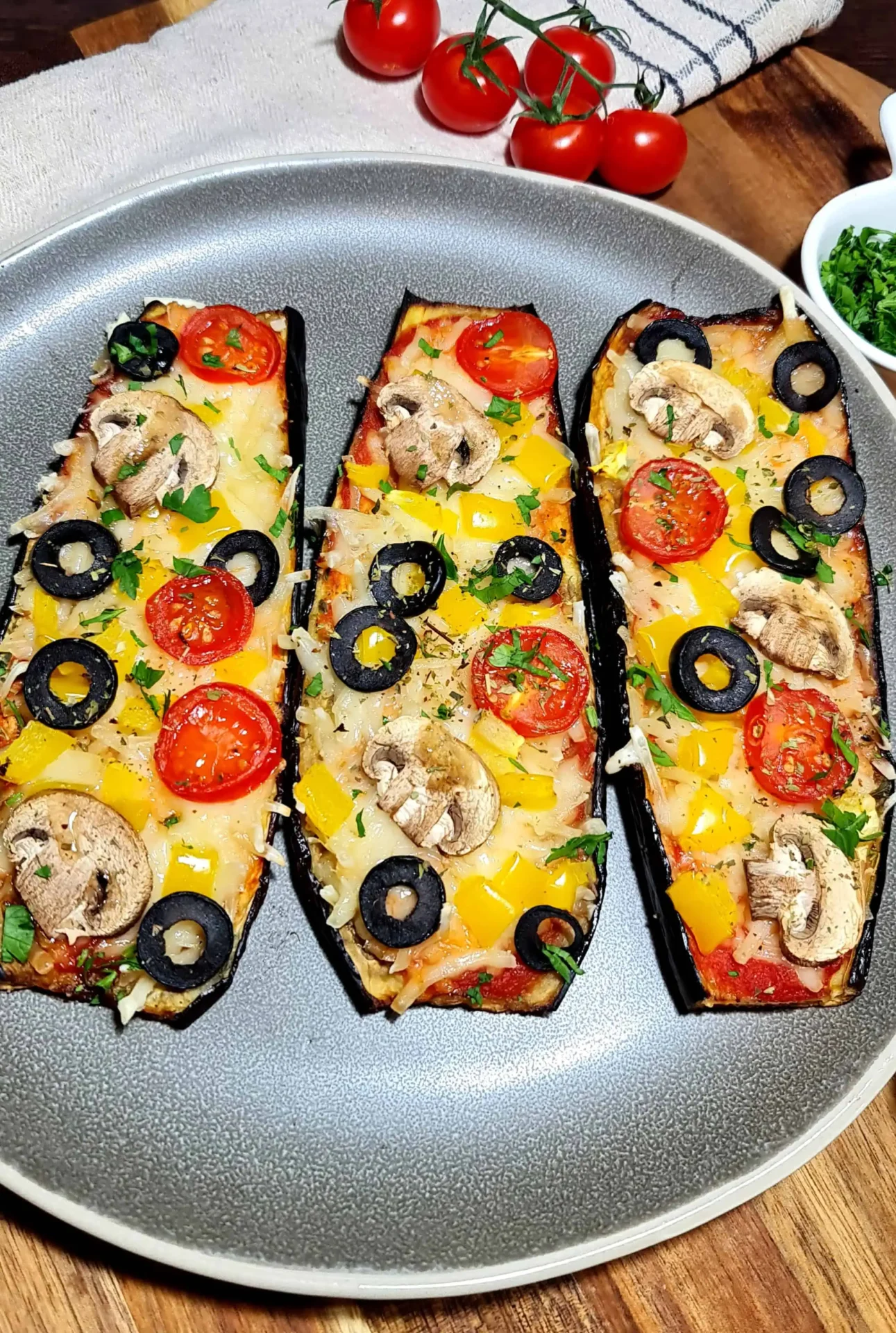 I could eat these delicious eggplants every day! Very quick and easy❗️ Three slices of eggplant prepared as mini pizzas, each topped with cherry tomato halves, black olive slices, yellow bell pepper pieces, mushroom slices, shredded cheese, and chopped parsley, arranged on a gray plate with a background of fresh cherry tomatoes on the vine, a dish with chopped herbs, and a striped cloth napkin.