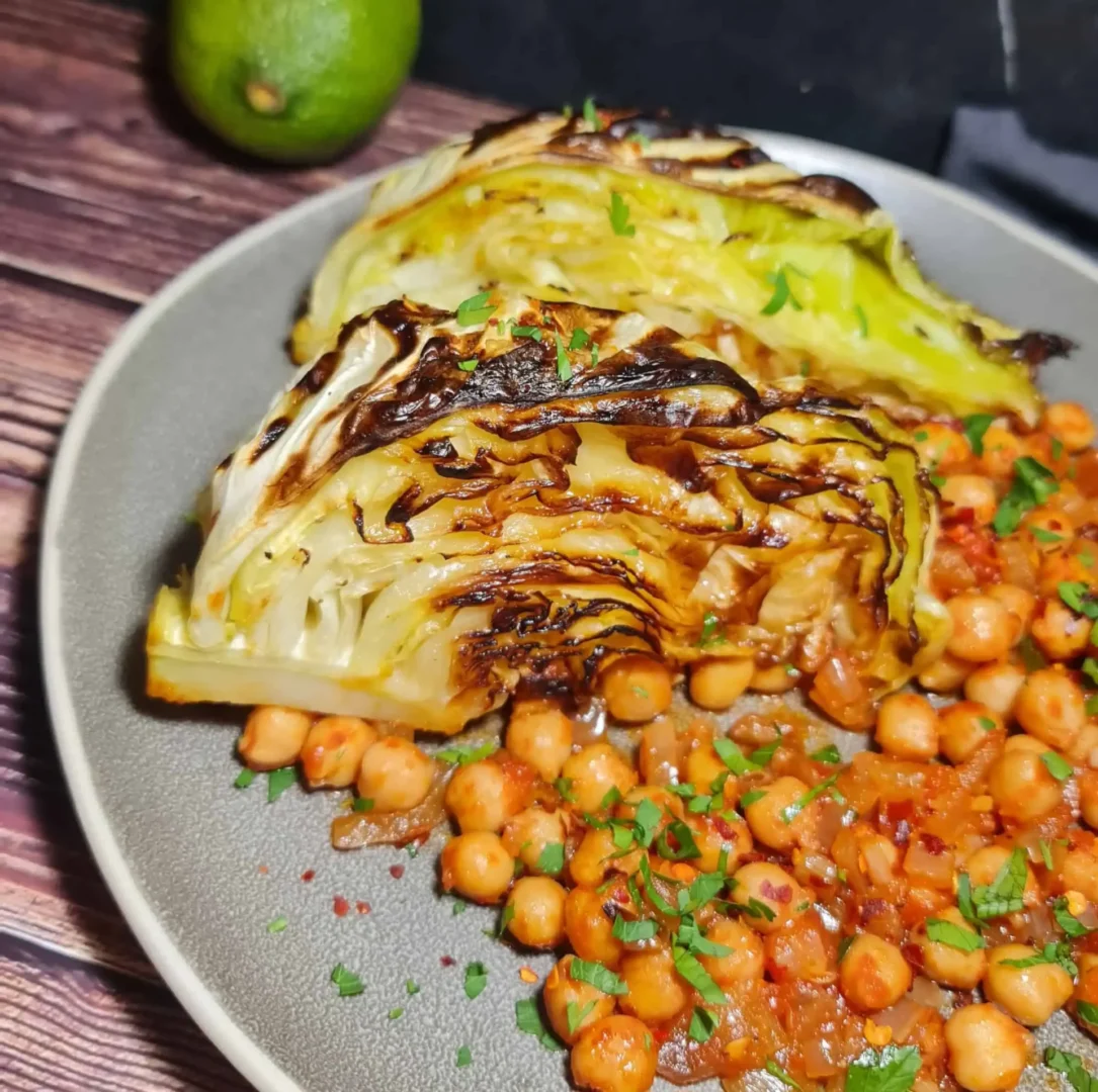 A dish of roasted cabbage wedges garnished with fresh herbs, served with chickpeas in a tomato-based sauce on a grey plate, with a lime and additional fresh herbs in the background.