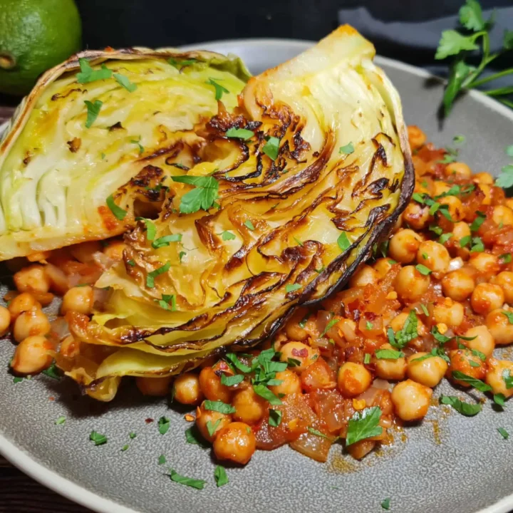 A dish of roasted cabbage wedges garnished with fresh herbs, served with chickpeas in a tomato-based sauce on a grey plate, with a lime and additional fresh herbs in the background.