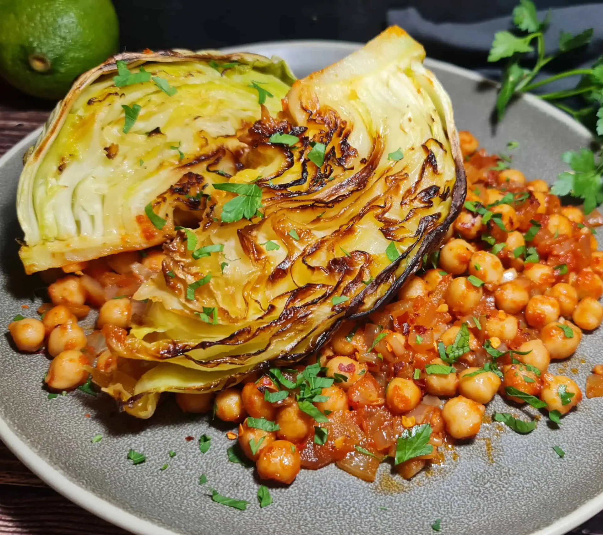 A dish of roasted cabbage wedges garnished with fresh herbs, served with chickpeas in a tomato-based sauce on a grey plate, with a lime and additional fresh herbs in the background.