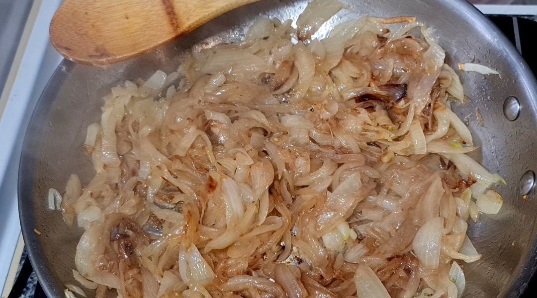 Caramelized onions in a stainless steel pan with a wooden spatula, prepared for a vegan French onion potato bake.