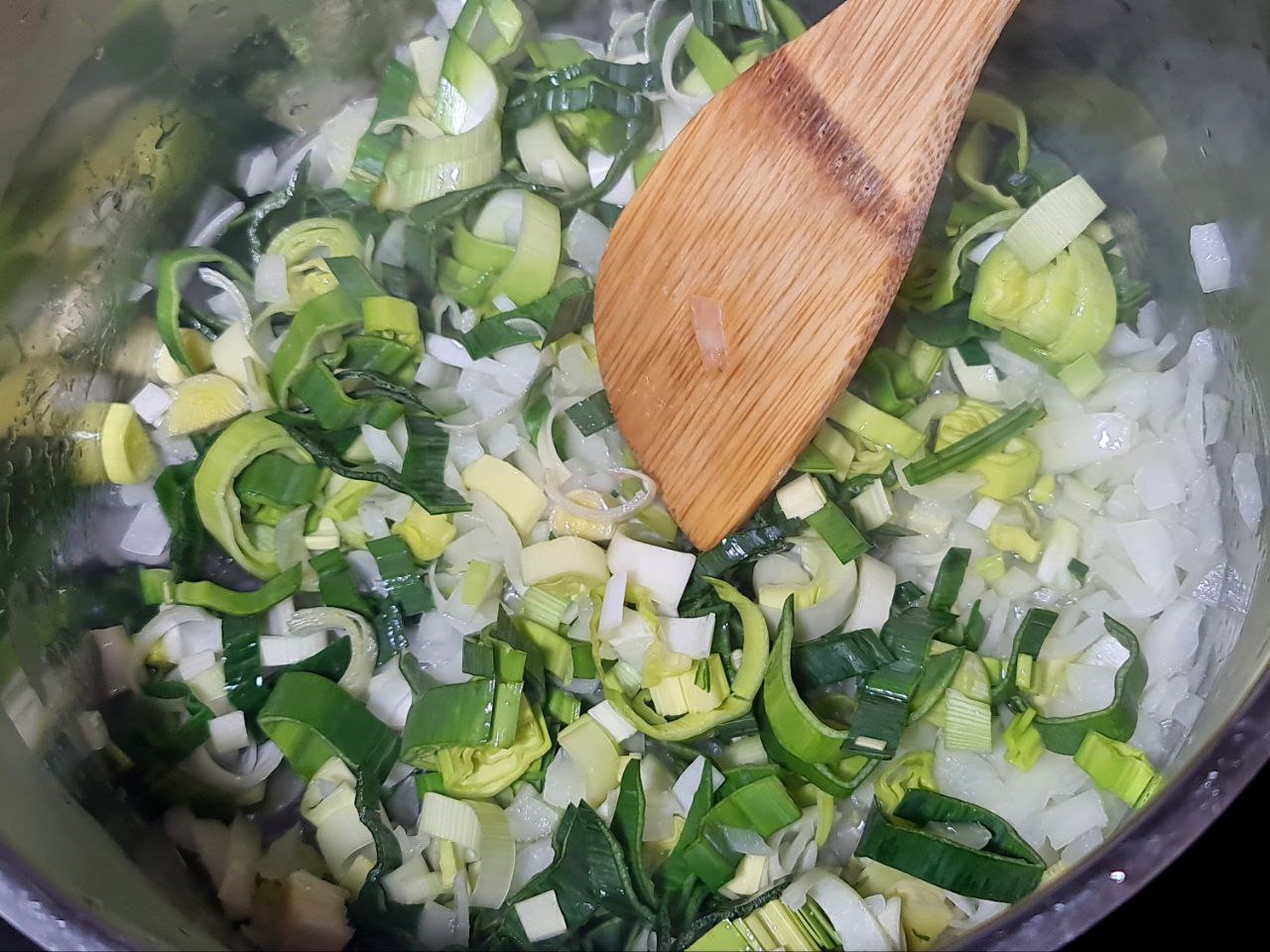 Chopped leeks and onions being sautéed in a pot with a wooden spoon.