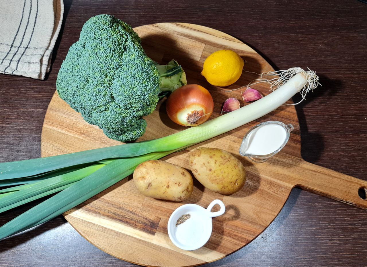 Ingredients for creamy vegan broccoli soup including broccoli, potatoes, leek, onion, garlic, lemon, salt, pepper, and plant-based milk on a wooden cutting board.