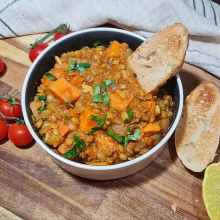 A bowl of sweet potato lentil soup garnished with fresh herbs, accompanied by toasted bread slices, cherry tomatoes, and a lemon half on a wooden surface.