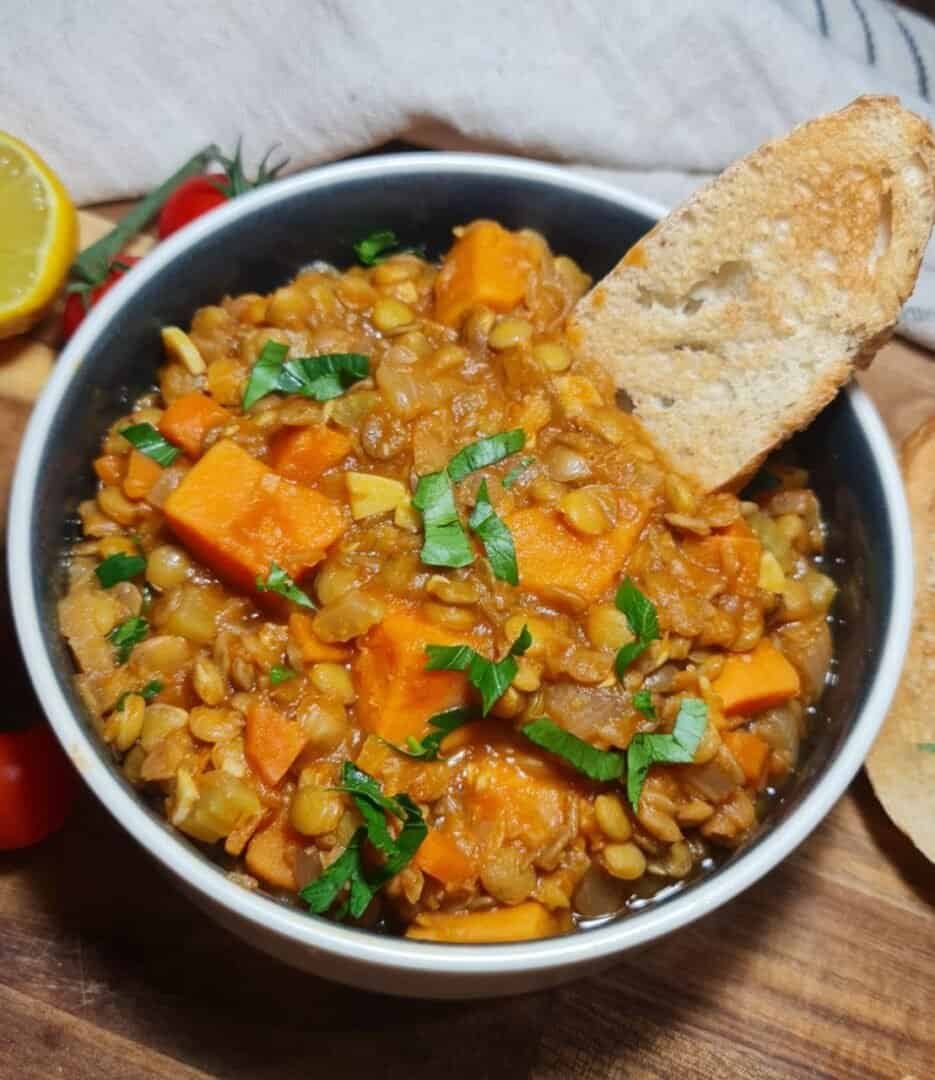 A bowl of sweet potato lentil soup garnished with fresh herbs, accompanied by toasted bread slices, cherry tomatoes, and a lemon half.