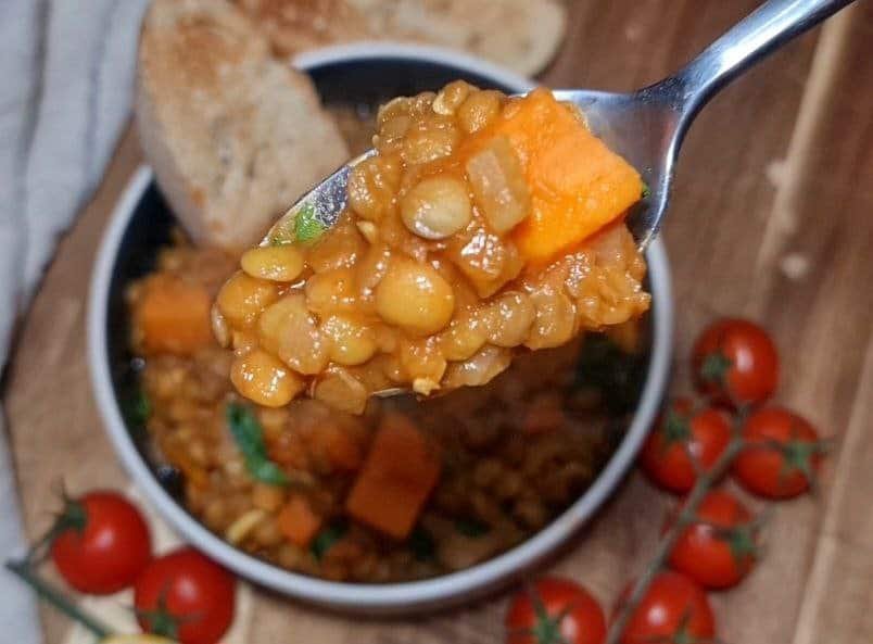 A close-up of a spoonful of sweet potato lentil soup with visible chunks of sweet potato, lentils, and herbs, with a bowl of the soup and cherry tomatoes in the background.