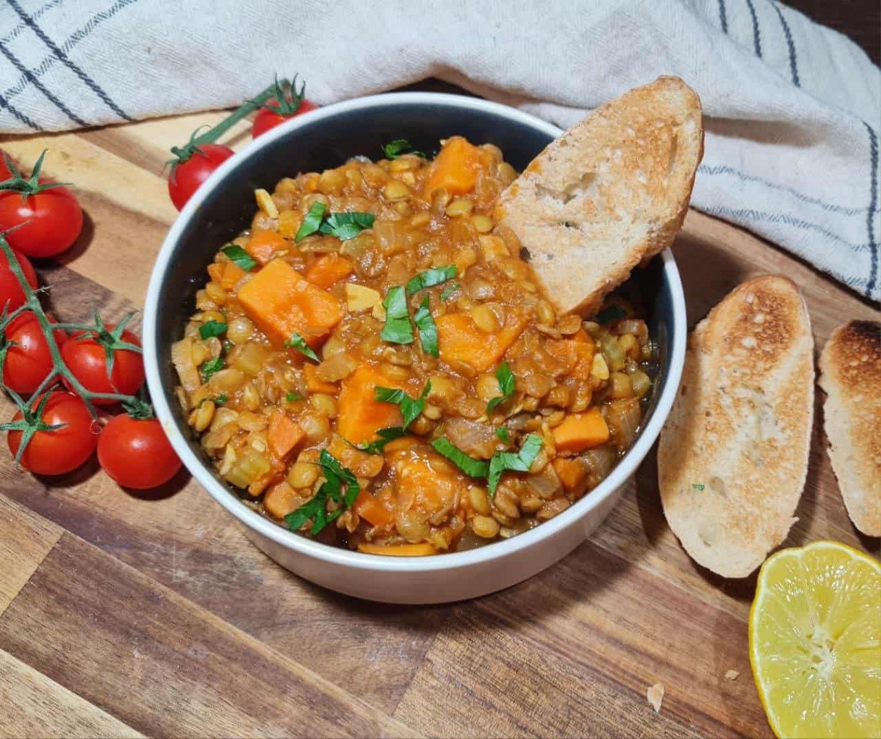 A bowl of sweet potato lentil soup garnished with fresh herbs, accompanied by toasted bread slices, cherry tomatoes, and a lemon half on a wooden surface.