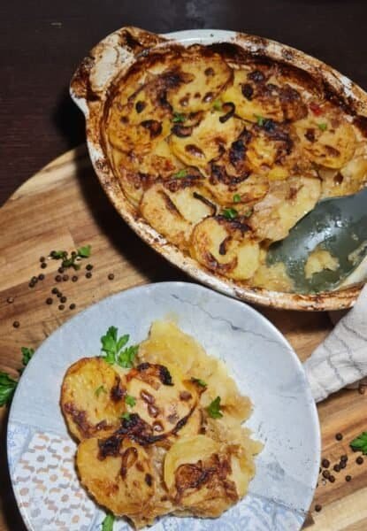 A baked dish of vegan French onion potato bake with a serving on a plate, garnished with fresh parsley.