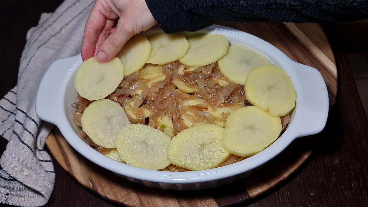 Hand placing sliced potatoes on top of caramelized onions in a white baking dish for a vegan French onion potato bake.