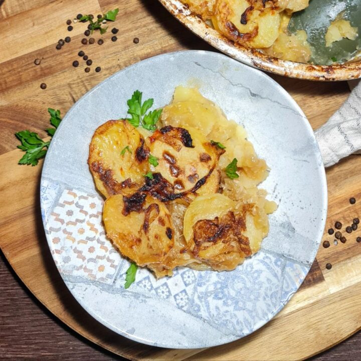 A plate of vegan French onion potato bake garnished with fresh parsley, served on a wooden board with peppercorns scattered around.