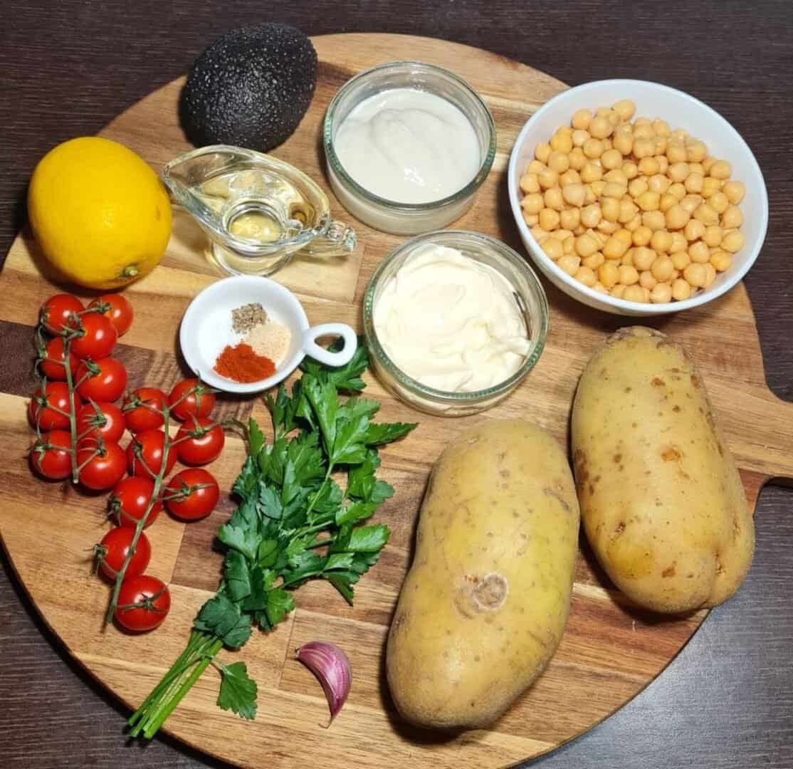  Ingredients for vegan kumpir including potatoes, chickpeas, cherry tomatoes, parsley, garlic, lemon, avocado, spices, oil, and sauces on a wooden board.