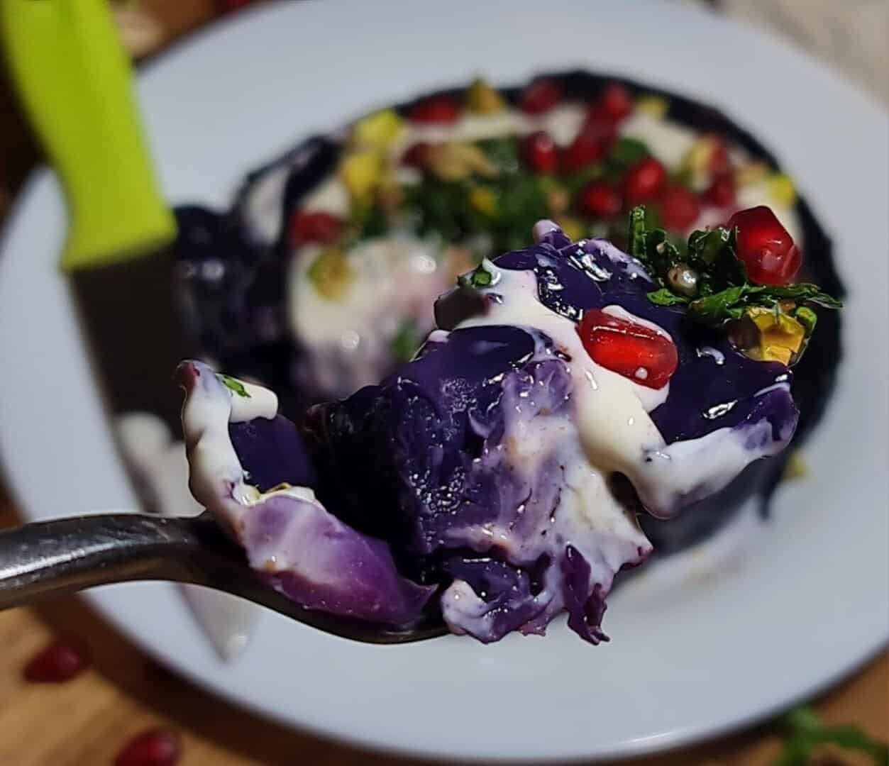 Close-up of a fork holding a piece of vegan roasted cabbage steak topped with creamy sauce, pomegranate seeds, and herbs.