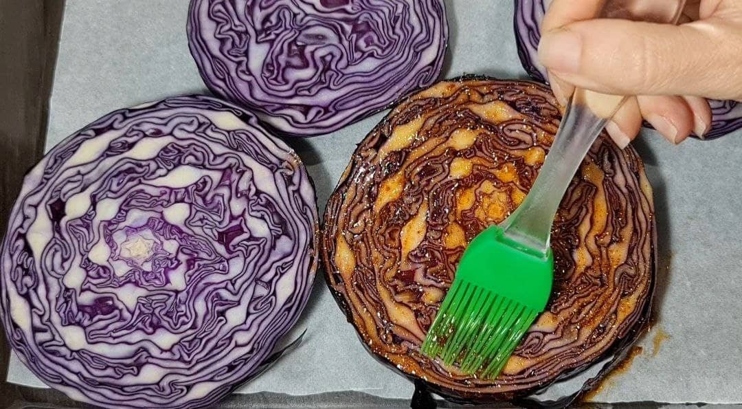 Hand brushing marinade on sliced purple cabbage steaks on a baking sheet.