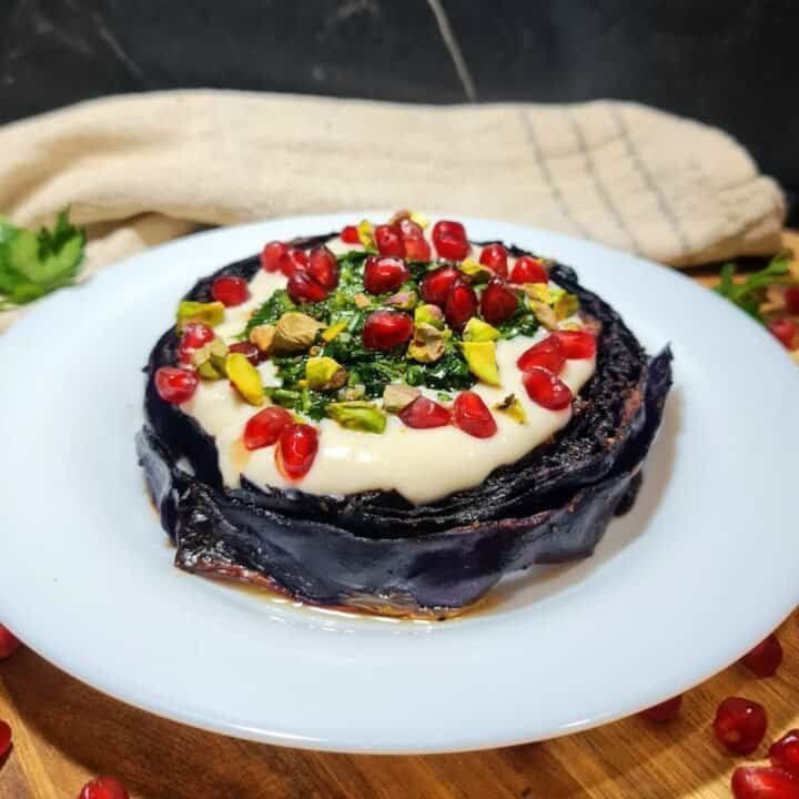 Vegan roasted red cabbage steak topped with creamy sauce, pomegranate seeds, chopped pistachios, and herbs on a white plate.