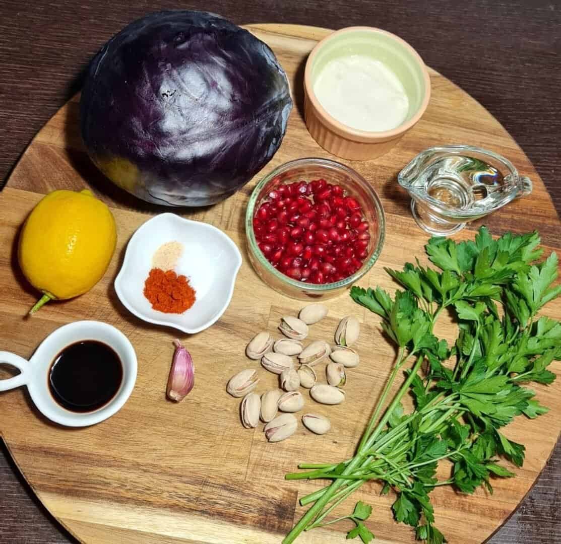 Ingredients for vegan roasted red cabbage steaks including red cabbage, lemon, pomegranate seeds, parsley, pistachios, garlic, spices, soy sauce, and a creamy dressing.