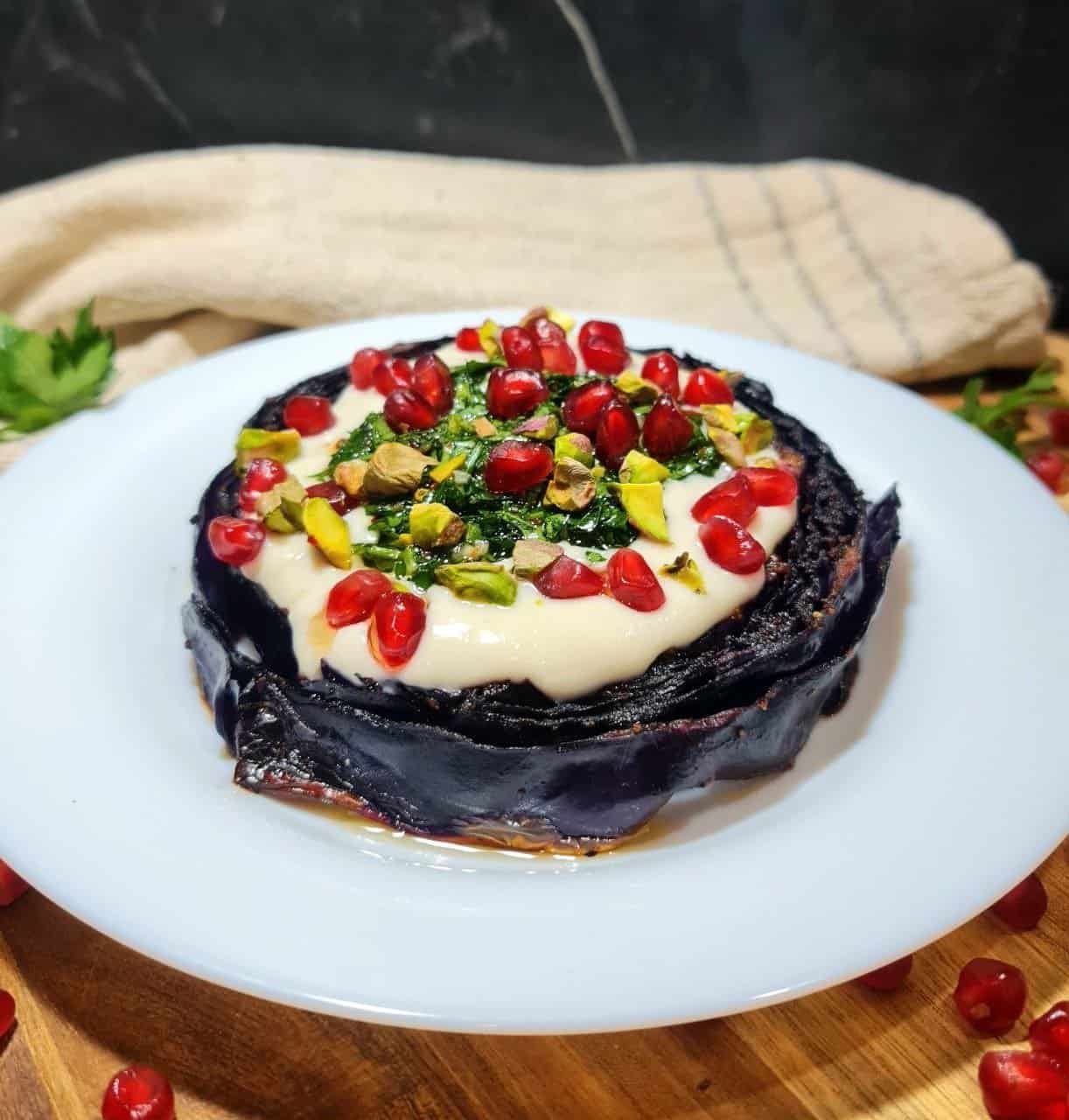 Vegan roasted red cabbage steak topped with creamy sauce, pomegranate seeds, chopped pistachios, and herbs on a white plate.