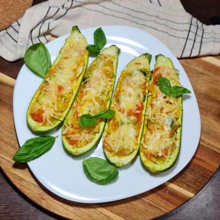 Four vegan stuffed zucchini boats topped with melted cheese and garnished with fresh basil leaves on a white plate.