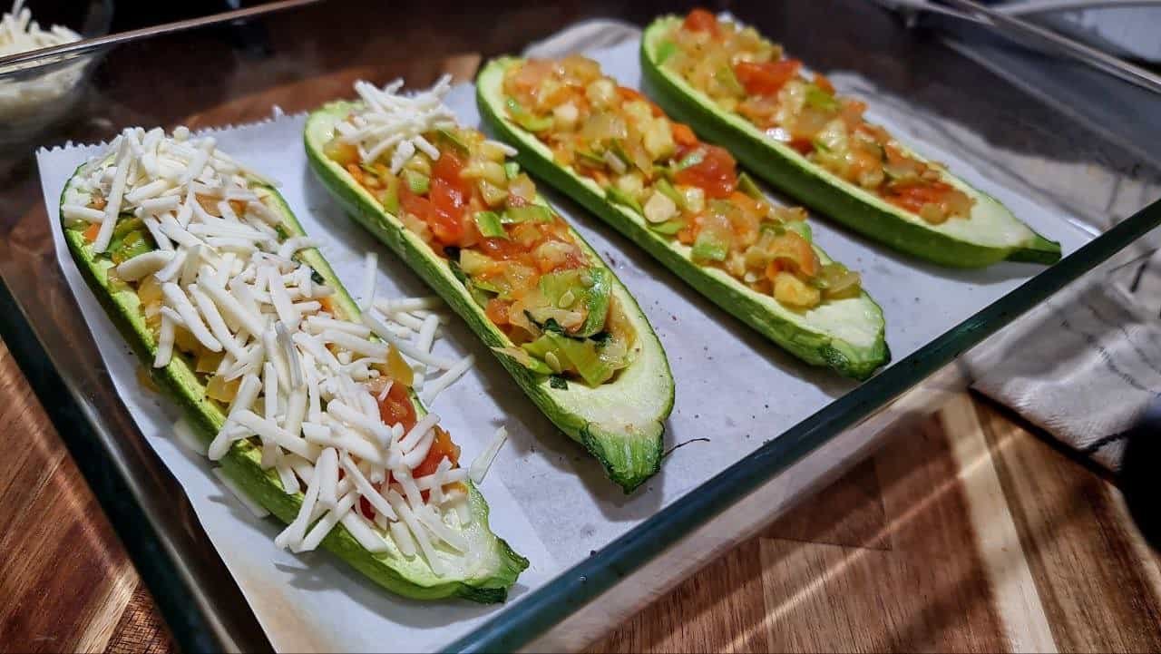 Four vegan stuffed zucchini halves filled with a colorful mixture of vegetables and topped with shredded vegan cheese, arranged in a glass baking dish.