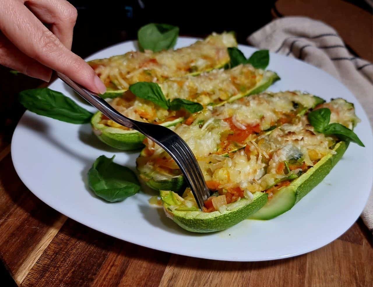 A plate of vegan stuffed zucchini boats garnished with fresh basil leaves, with a hand holding a fork ready to take a bite.