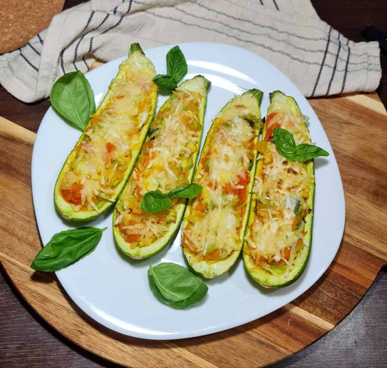 Four vegan stuffed zucchini boats topped with melted cheese and garnished with fresh basil leaves on a white plate.