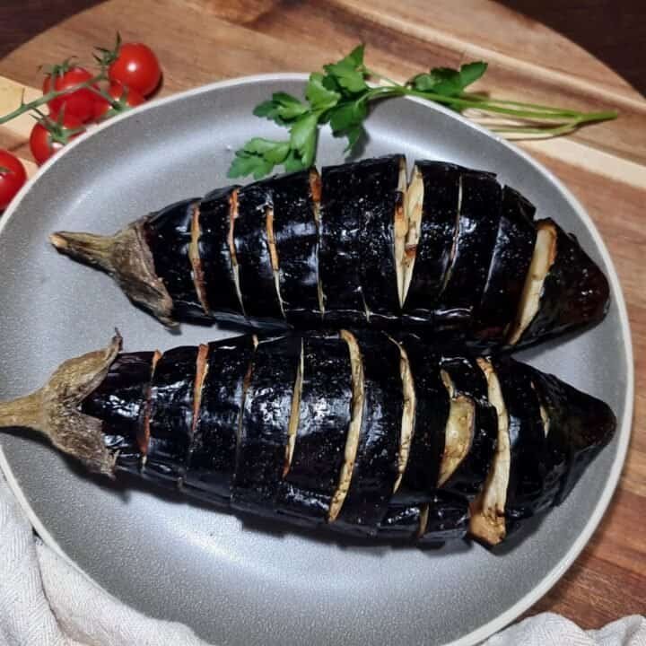 Two whole roasted eggplants sliced and served on a gray plate, with cherry tomatoes and a lemon wedge in the background.