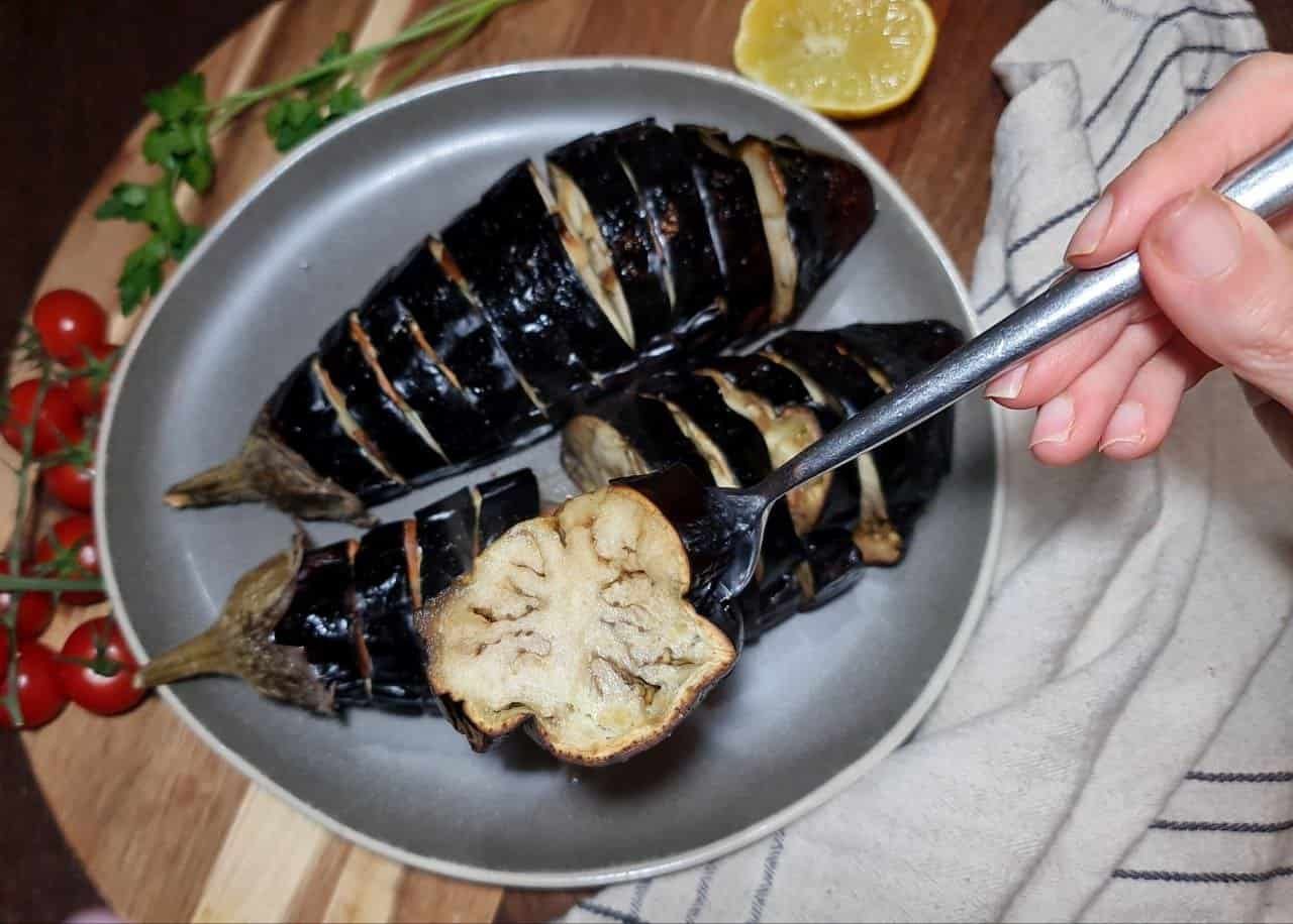 Whole roasted eggplant sliced and served on a plate with a fork holding a piece, garnished with cherry tomatoes, parsley, and a lemon half.