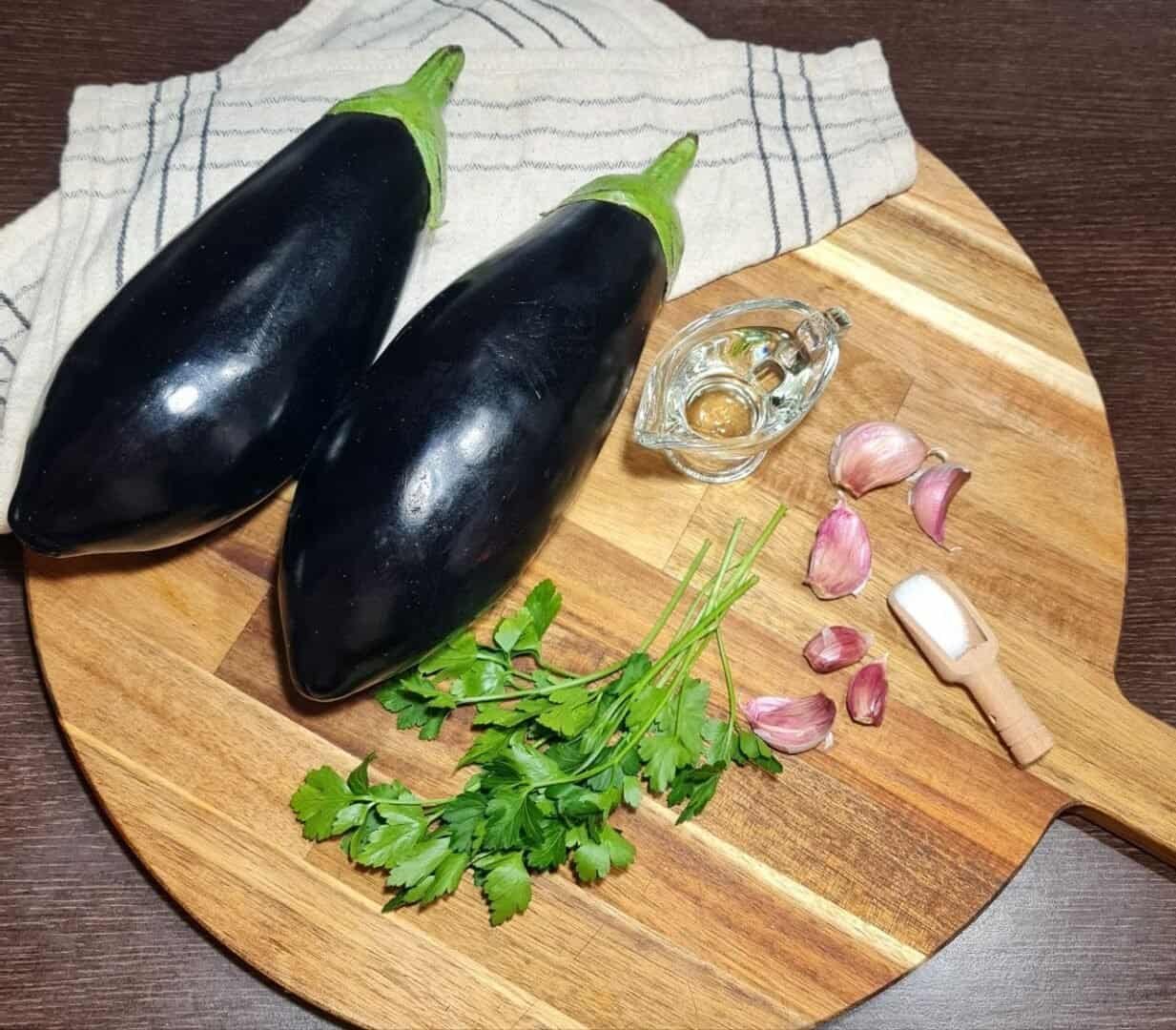 Ingredients for whole roasted eggplant including two eggplants, garlic cloves, parsley, salt, and oil on a wooden cutting board.