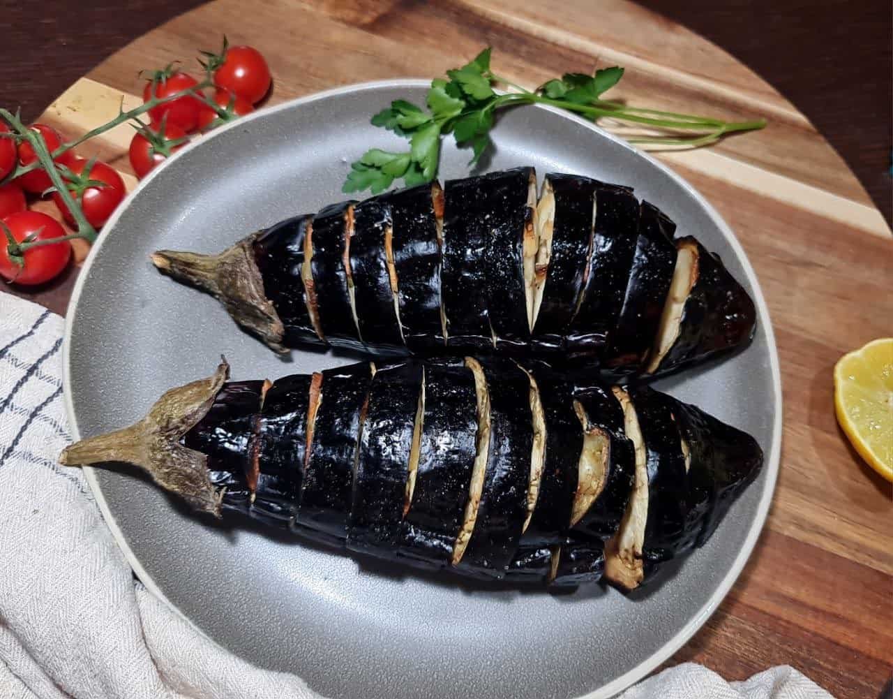 Two whole roasted eggplants sliced and served on a gray plate, with cherry tomatoes and a lemon wedge in the background.