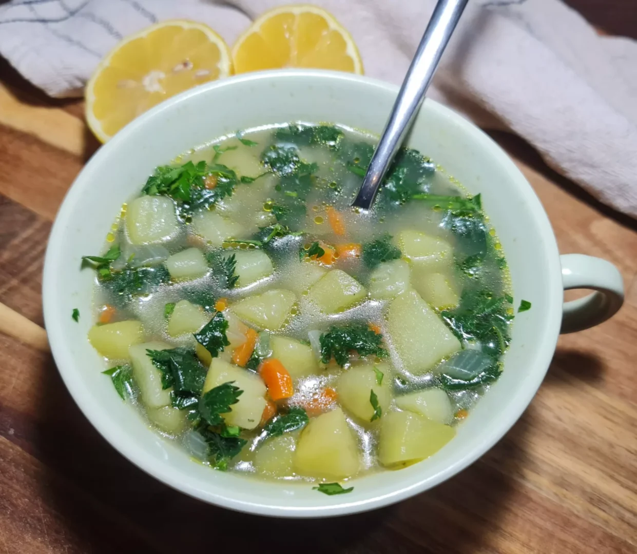 A bowl of Hearty Nettle Soup containing diced potatoes, carrots, onions, and parsley in a clear broth, garnished with two lemon halves in the background on a rustic cloth.