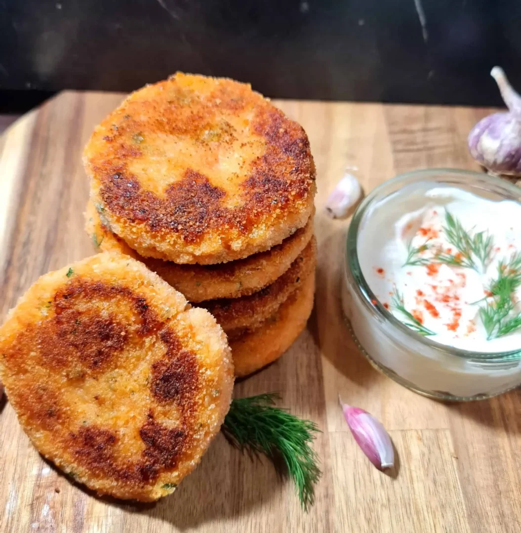 A stack of golden-brown Vegan Red Lentil Sweet Potato Patties on a wooden cutting board, accompanied by a small glass bowl of creamy garlic dip garnished with fresh dill and red paprika, with garlic cloves and a sprig of dill for decoration.