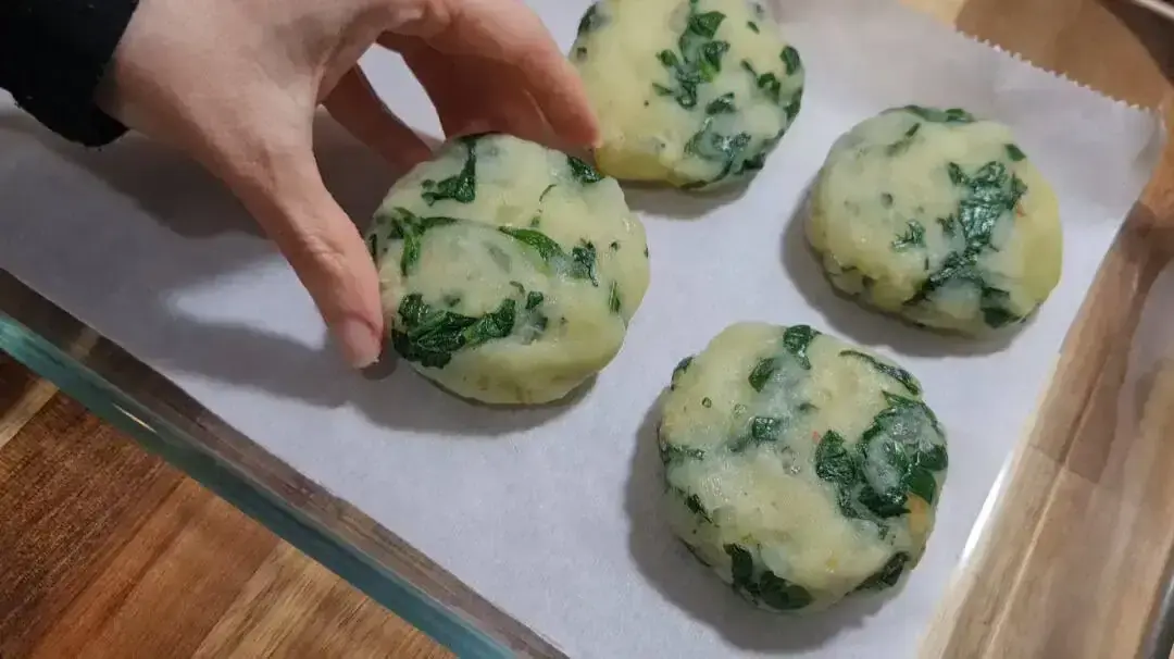 Four uncooked patties with visible green leafy vegetables, placed on parchment paper. A hand is holding one patty, showcasing the preparation process for Walnut Maple Sweet Potato Rounds.