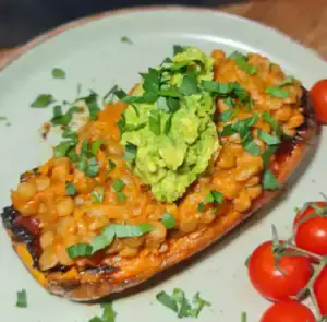 A roasted sweet potato half topped with a lentil mixture in sauce, garnished with chopped fresh cilantro and a dollop of mashed avocado, served with cherry tomatoes on the side.