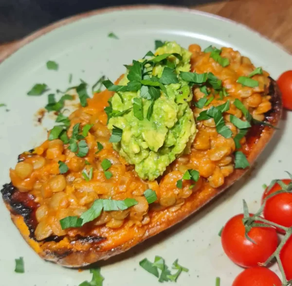 A roasted sweet potato half topped with a lentil mixture in sauce, garnished with chopped fresh cilantro and a dollop of mashed avocado, served with cherry tomatoes on the side.