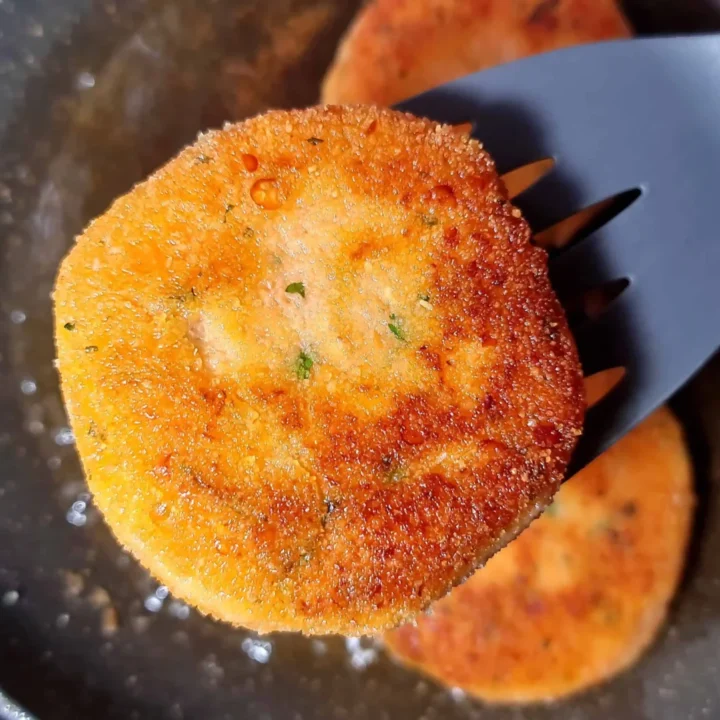 A close-up of a golden-brown Vegan Red Lentil Sweet Potato Patty held by a gray spatula over a frying pan, with another patty visible in the background.