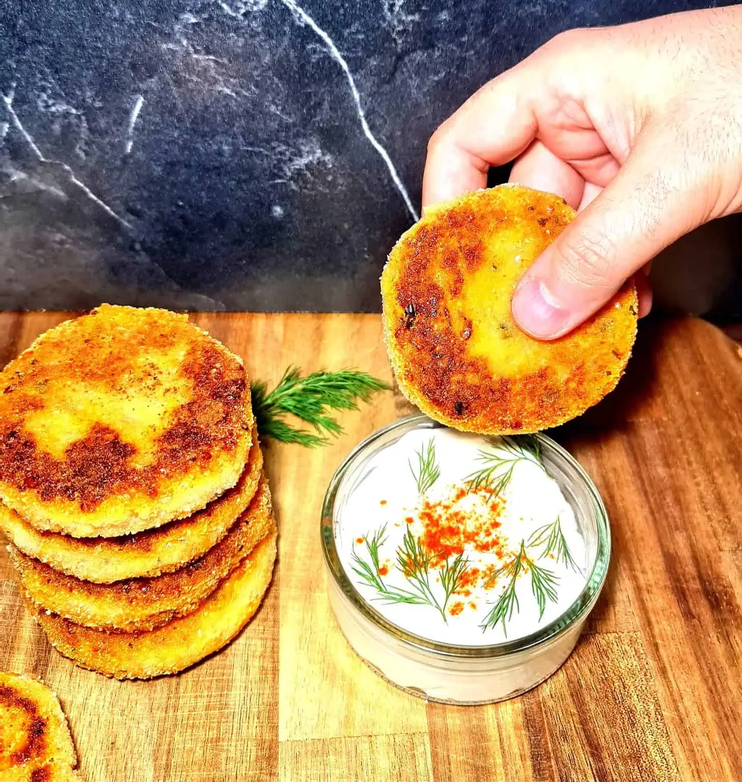 A golden-brown Vegan Red Lentil Sweet Potato Patty held above a small glass bowl filled with creamy white dip garnished with dill and red seasoning, alongside a stack of patties on a wooden surface.