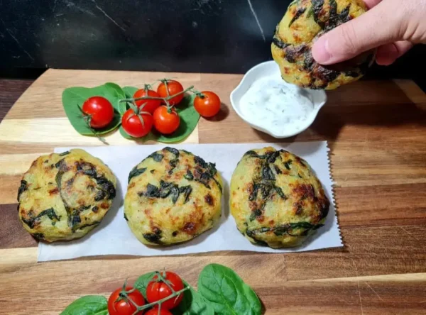 A hand dipping a golden-brown Walnut Maple Sweet Potato Round, with green bits visible, into a small white bowl of creamy herb sauce. Two additional rounds sit on parchment paper on a wooden surface, accompanied by cherry tomatoes and a green leaf.