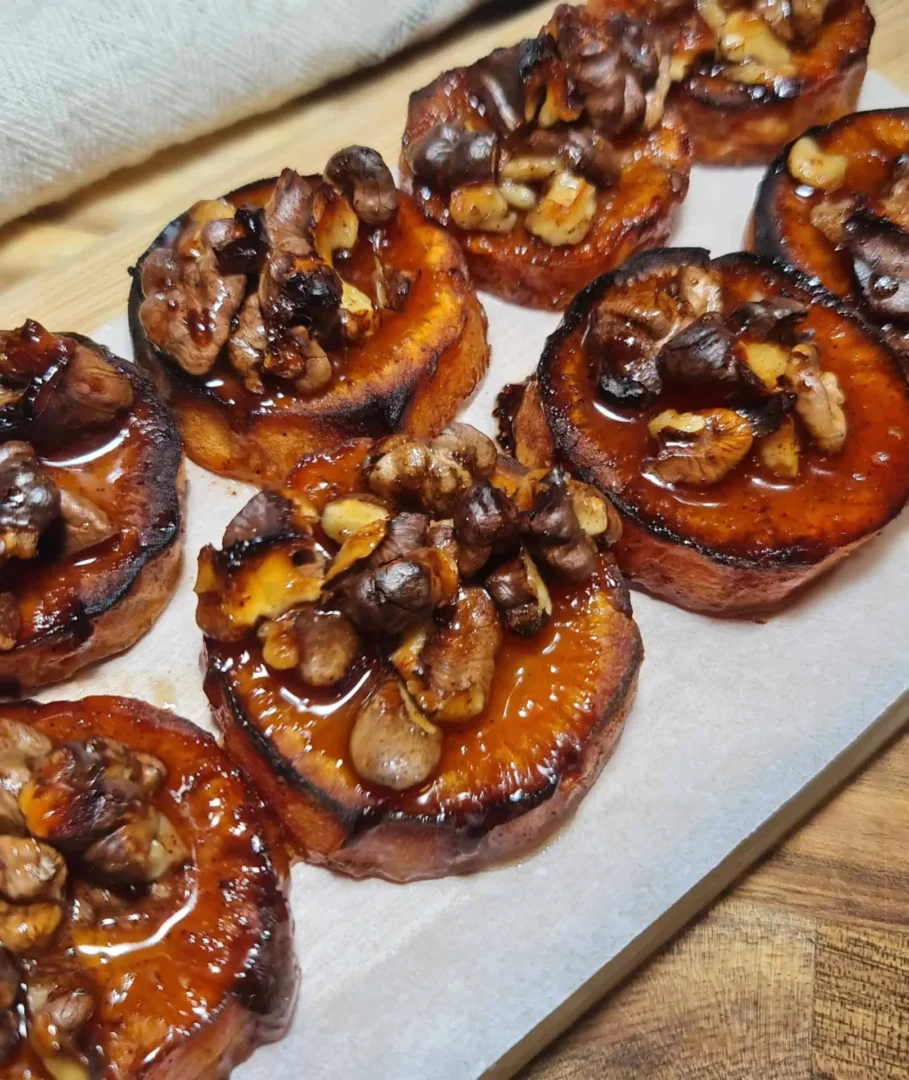 A display of roasted sweet potato rounds on parchment paper, each topped with a caramelized mixture of nuts coated in a glossy glaze, arranged on a wooden surface.