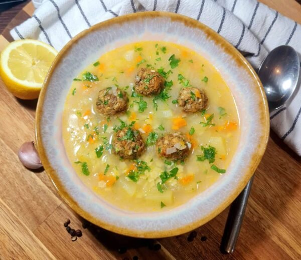 Rustic bowl of vegan lentil meatball soup topped with parsley, surrounded by lemon wedge, spoon, and striped napkin.