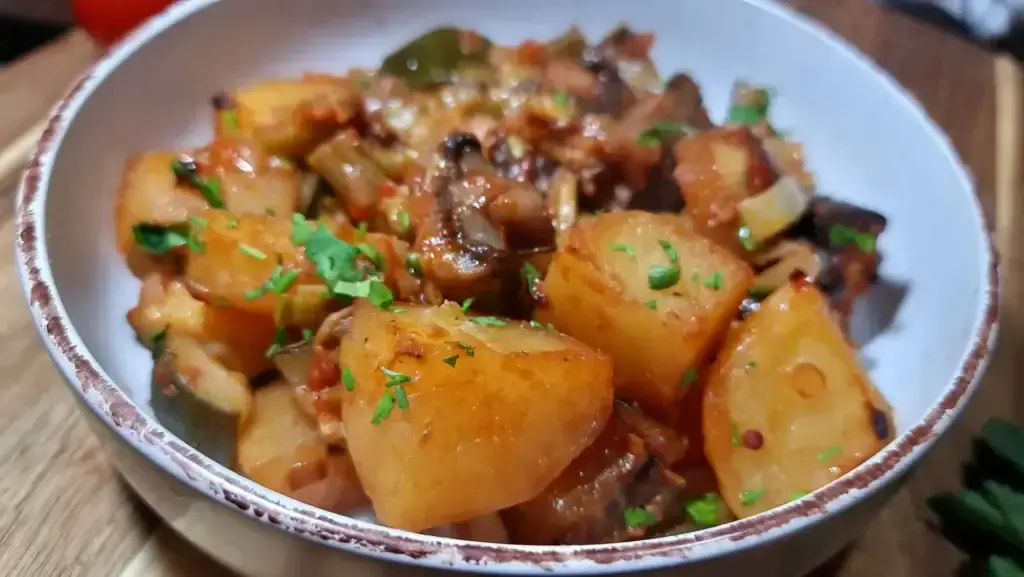 A bowl of Mediterranean Veggie Casserole, featuring cooked chunks of potatoes, mushrooms, zucchini, and other vegetables in a rich tomato-based sauce, garnished with fresh chopped herbs.