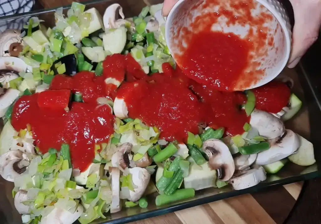 A glass baking dish filled with sliced mushrooms, chopped green beans, diced zucchini, and leeks being drizzled with a red tomato-based sauce from a white container.