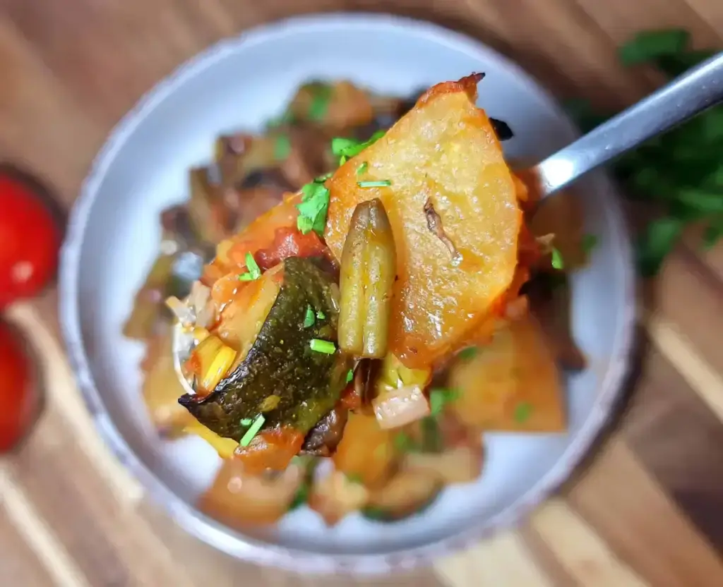 A close-up of a spoon holding Mediterranean Vegetable Casserole with chunks of zucchini, potatoes, and green beans in a tomato-based sauce, garnished with chopped herbs. A bowl of the casserole and fresh vegetables are visible in the blurred background.