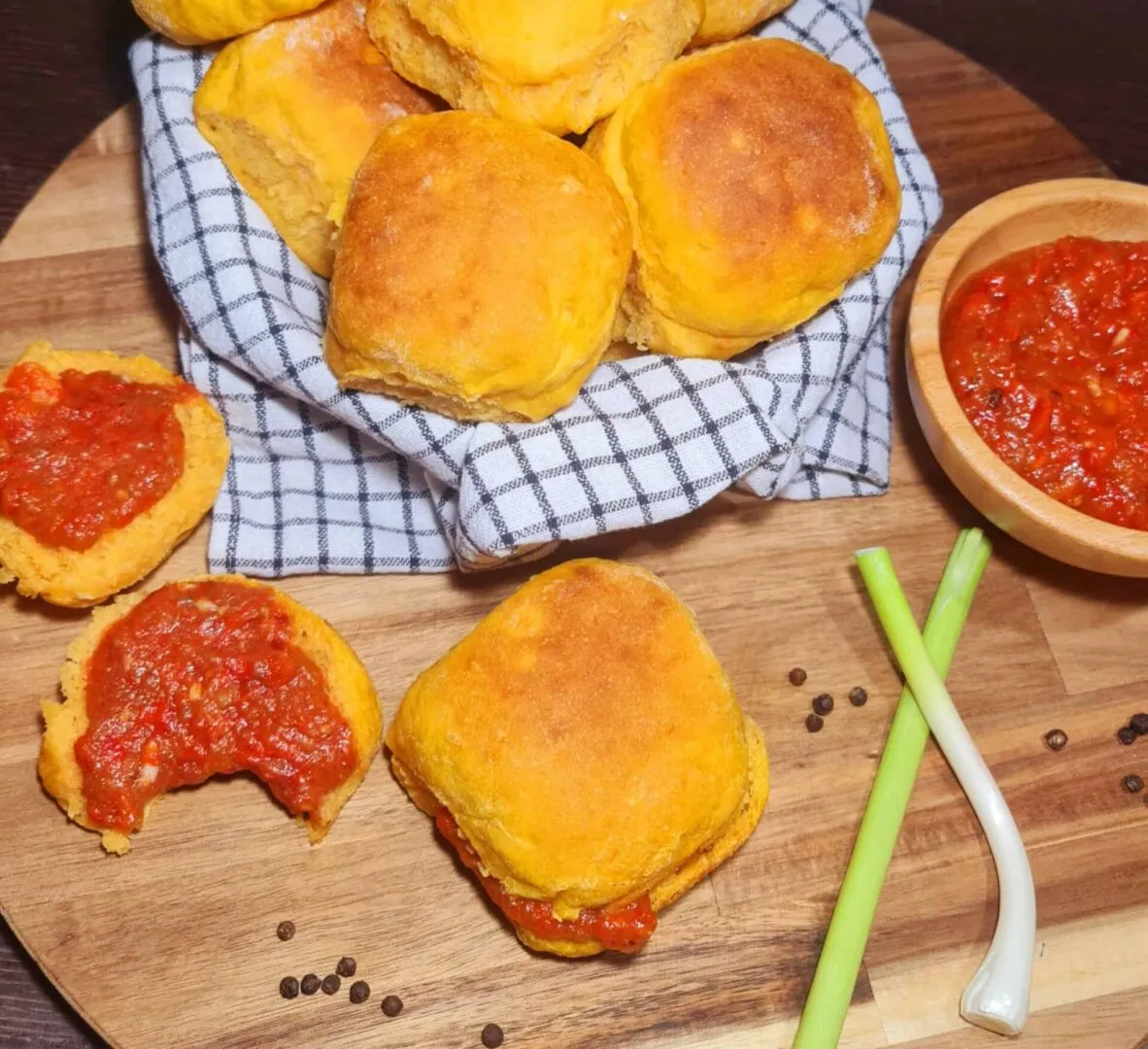 Basket lined with checkered cloth filled with golden Sweet Potato Dinner Rolls, surrounded by pepper sauce, spring onions, and peppercorns.